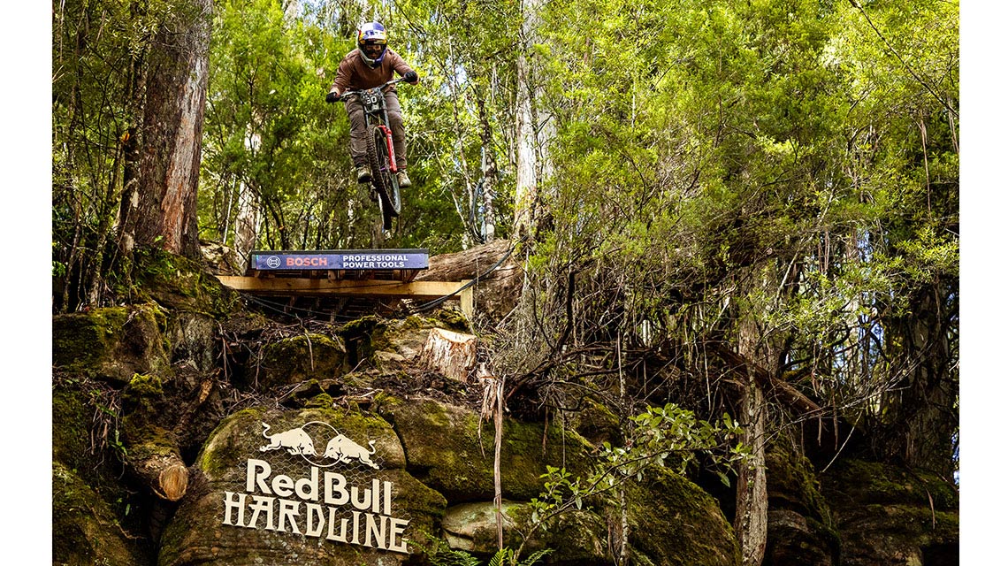 A cyclist jumps over a wooden ramp in the forest, Red Bull logo visible.
