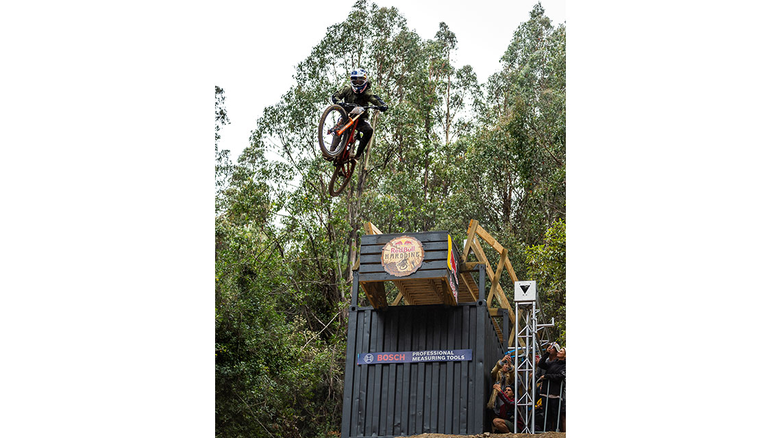 A mountain biker jumps from a wooden structure in a forest.