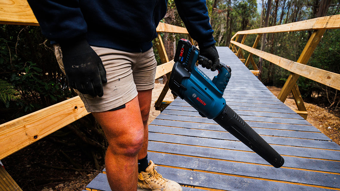 A craftsman holds a blue Bosch leaf blower on a wooden bridge.