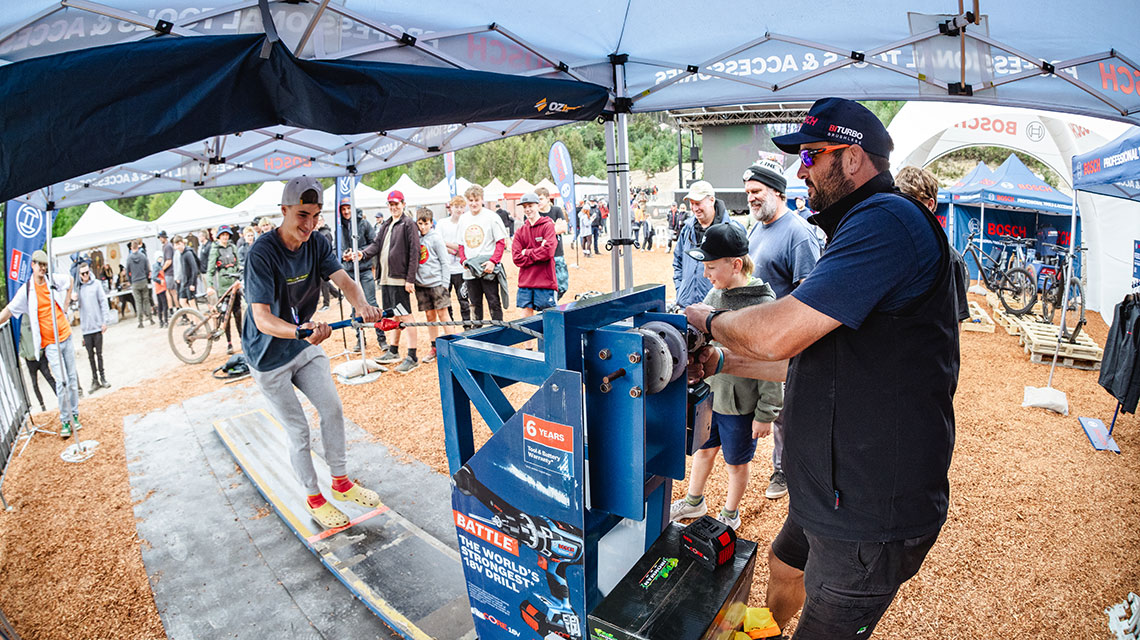 Man on skateboard tests Bosch tool, spectators in the background.