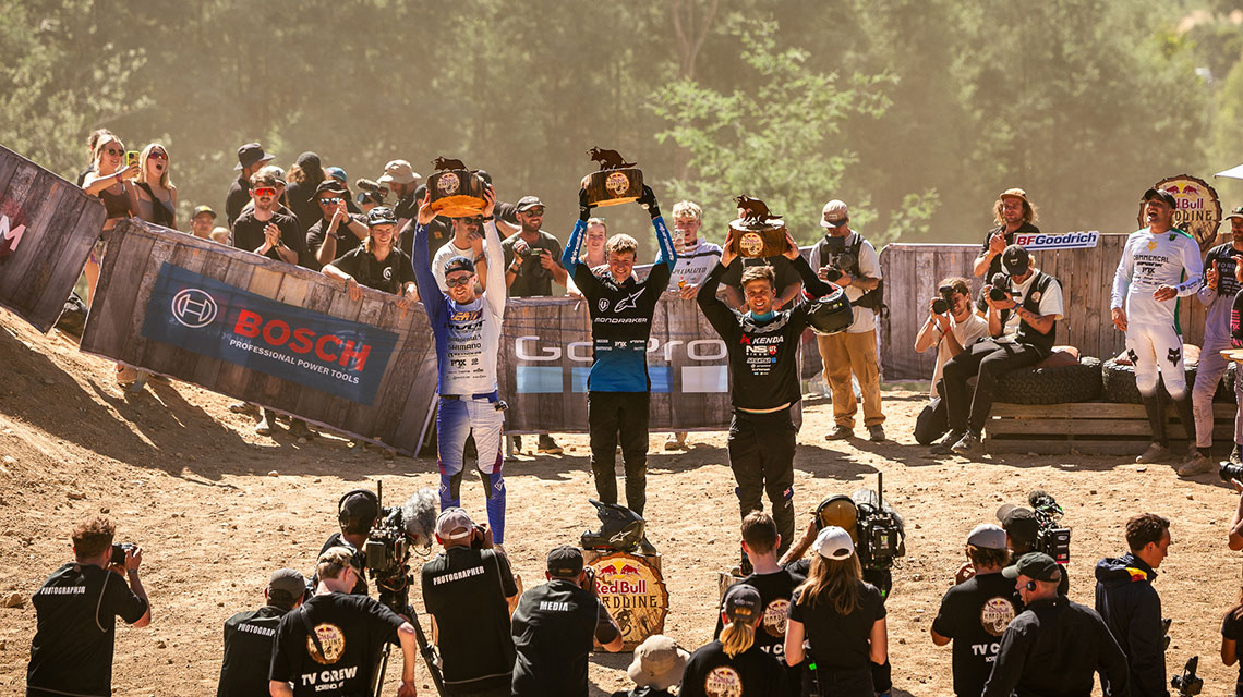 Three winners hold trophies on a dusty podium, spectators applaud.