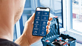 A man holds a smartphone with an app in his hand, suitcase in the background.