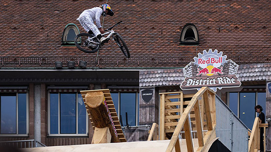 A cyclist in white sportswear jumps with his mountain bike over a wooden ramp. In the background, a building with a red "Red Bull District Ride" sign can be seen. The scene conveys dynamism and action.